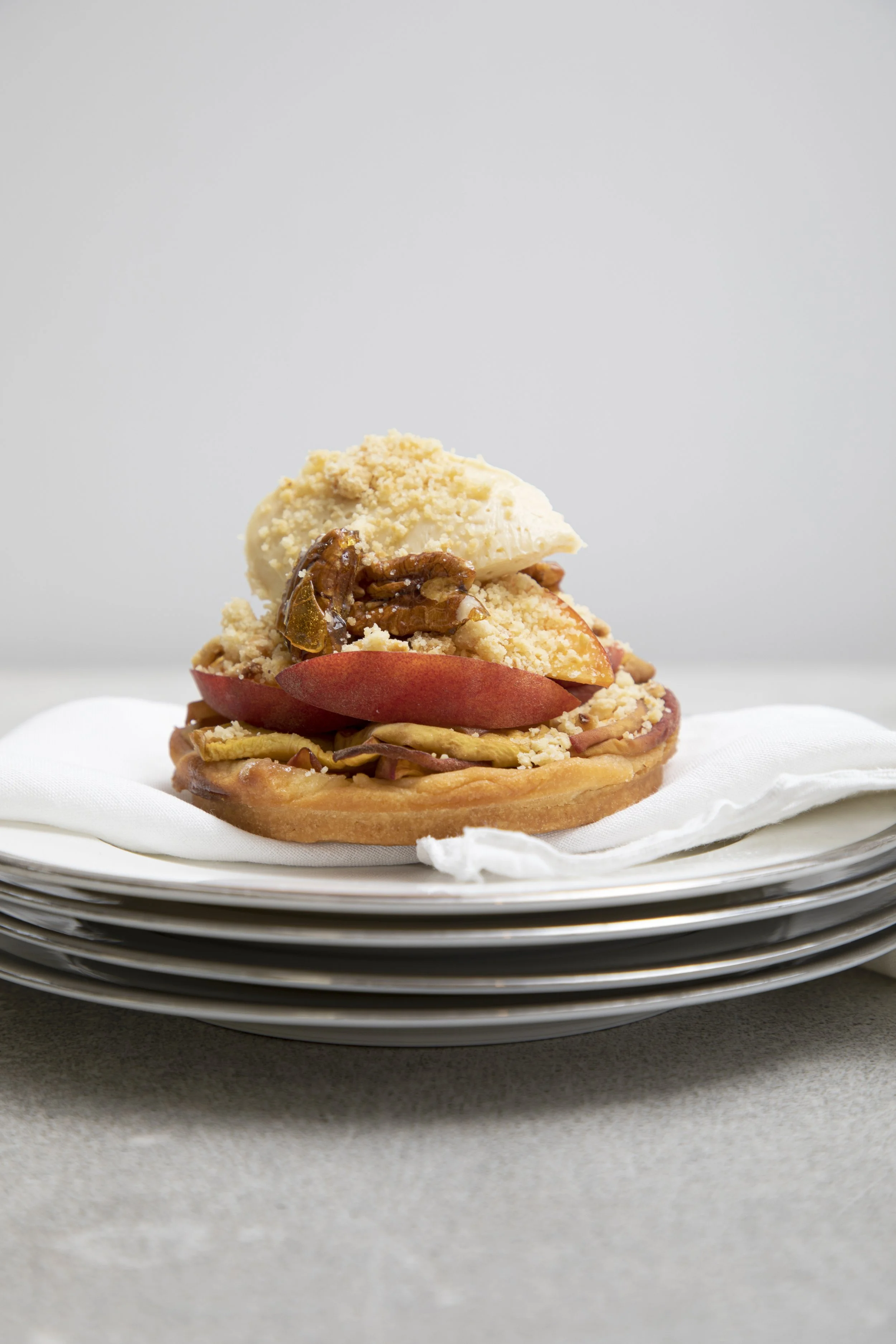 A close-up of a gourmet dessert pastry topped with slices of peach, chopped nuts, a scoop of vanilla ice cream, and drizzled caramel, presented on a white plate with a stack of similar plates underneath.
