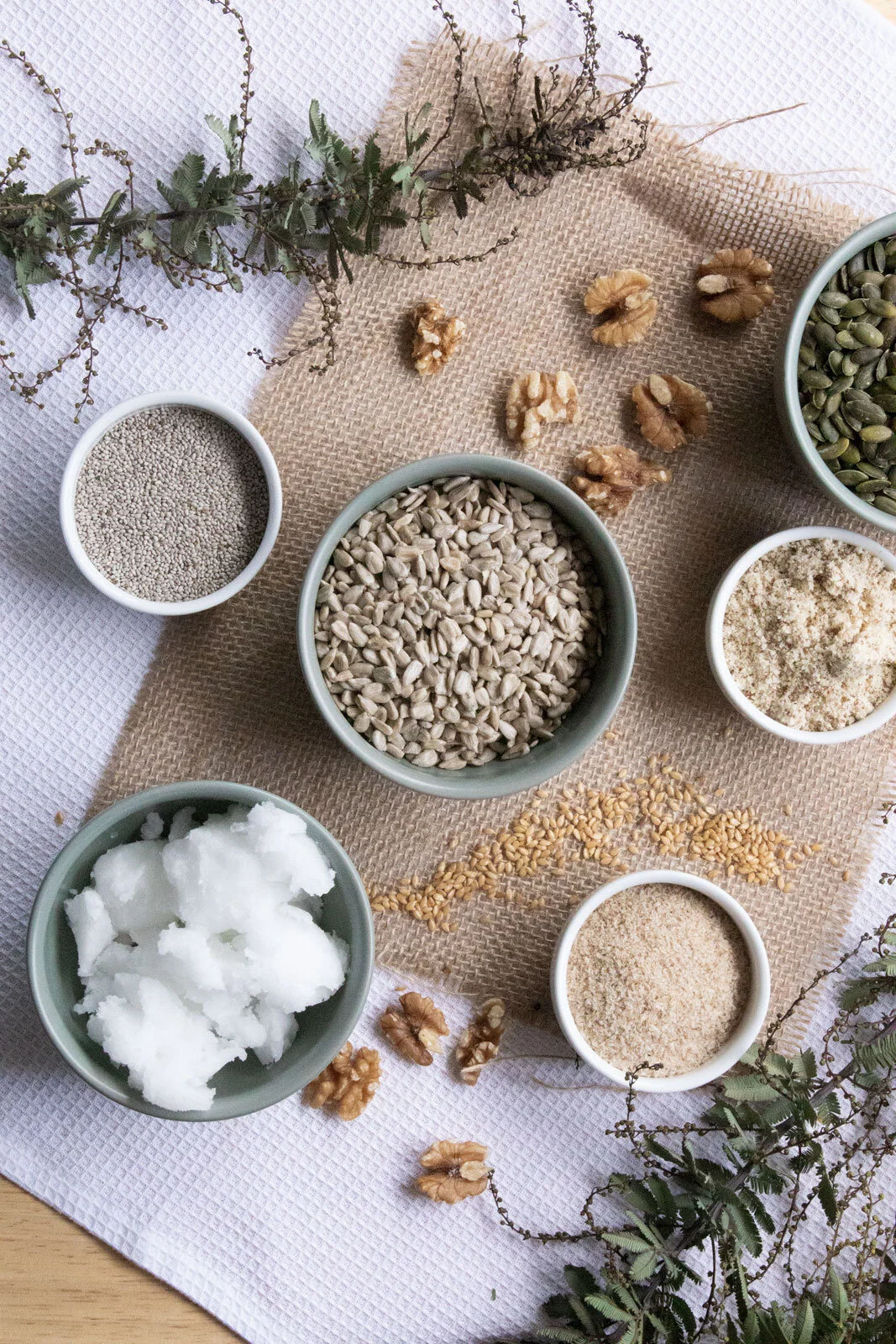 Assorted bowls of seeds, grains, nuts, and coconut flakes arranged on a beige and white textured tablecloth with sprigs of greenery.