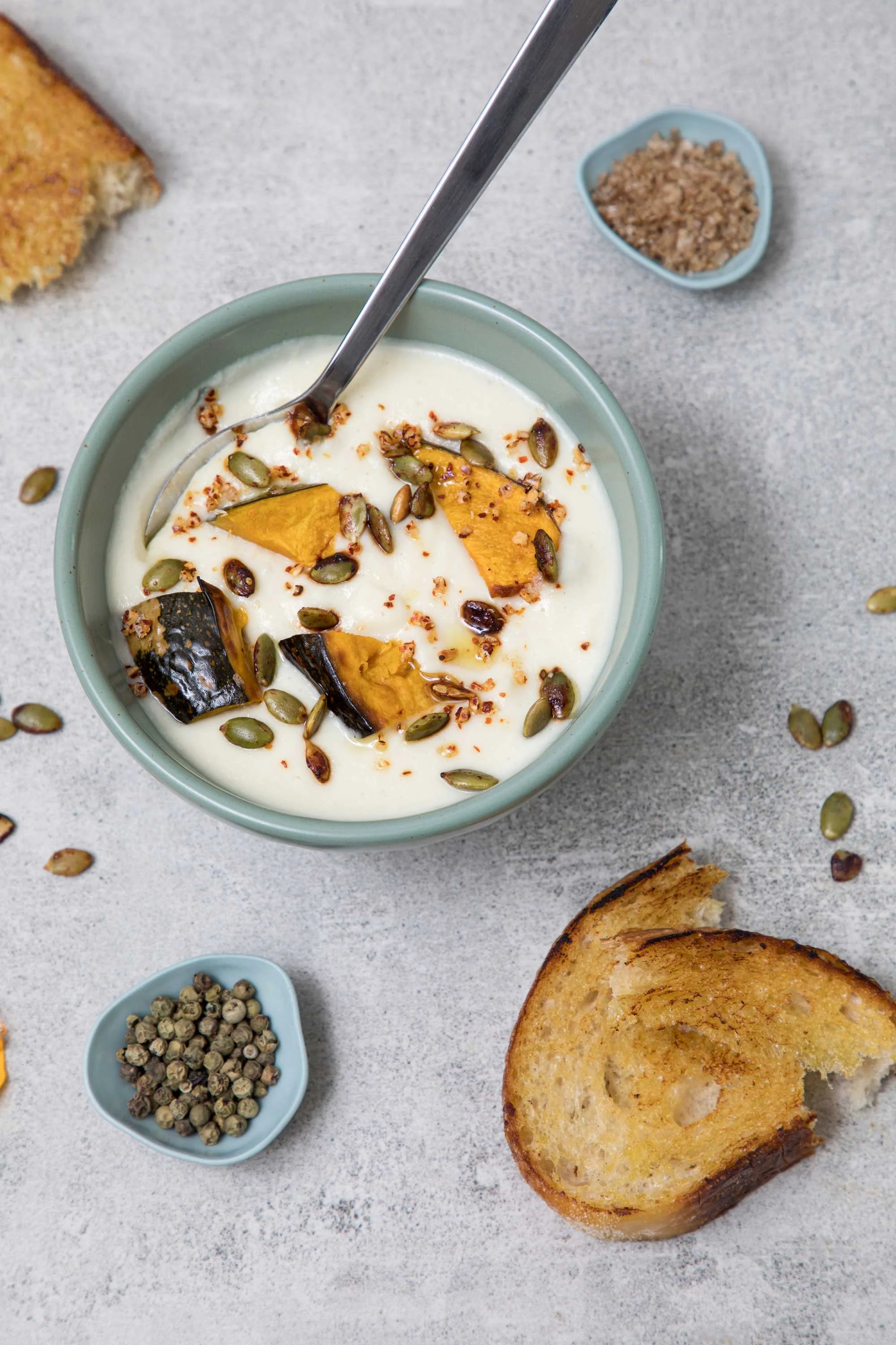 Bowl of creamy soup garnished with roasted pumpkin pieces and pumpkin seeds, with slices of toasted bread nearby on a light-colored surface.