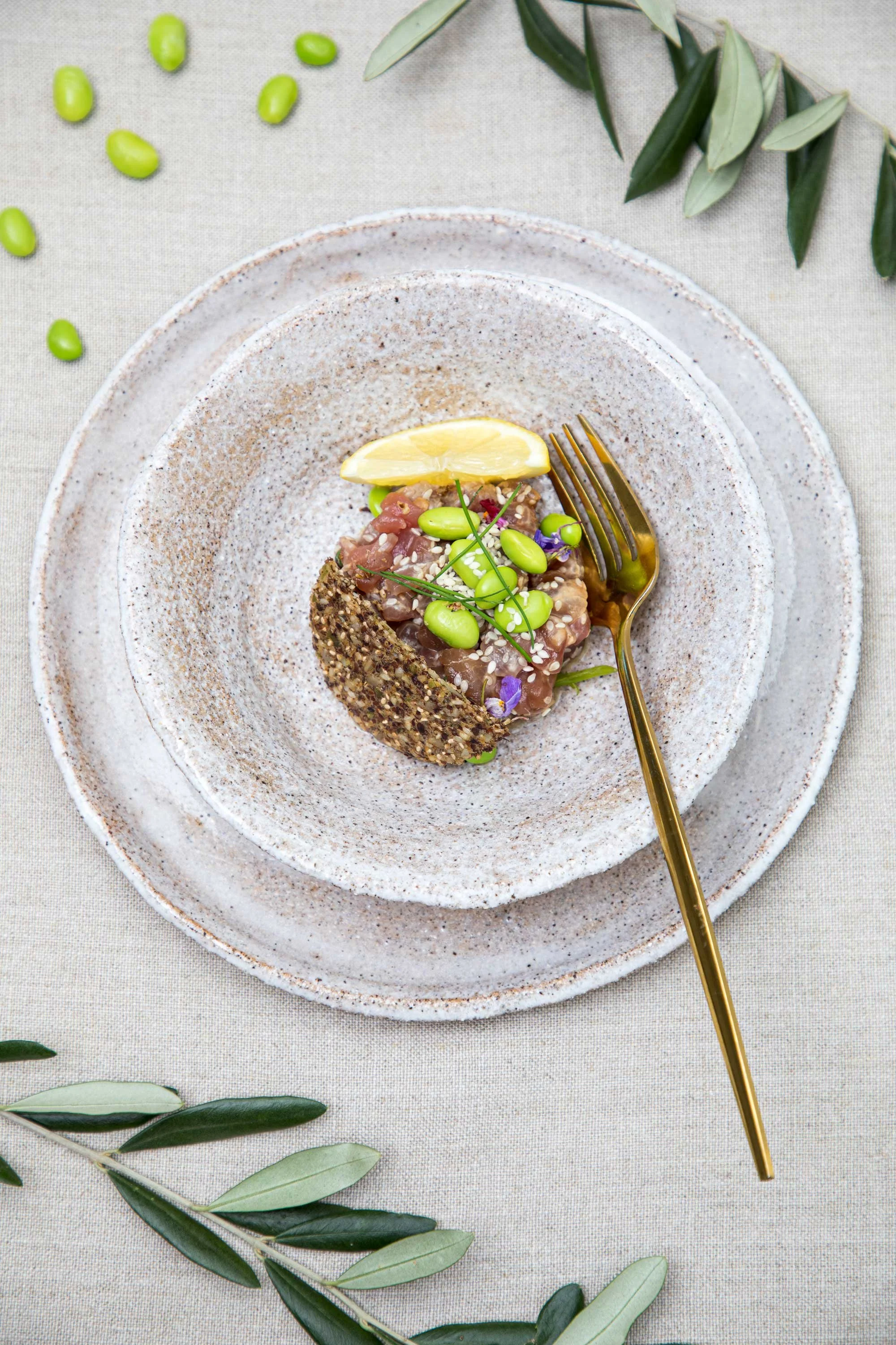 A plated serving of poke tuna with edamame, sesame seeds, and a lemon wedge. The dish is on a speckled ceramic plate with a gold fork, surrounded by green olive branches on a neutral cloth background.