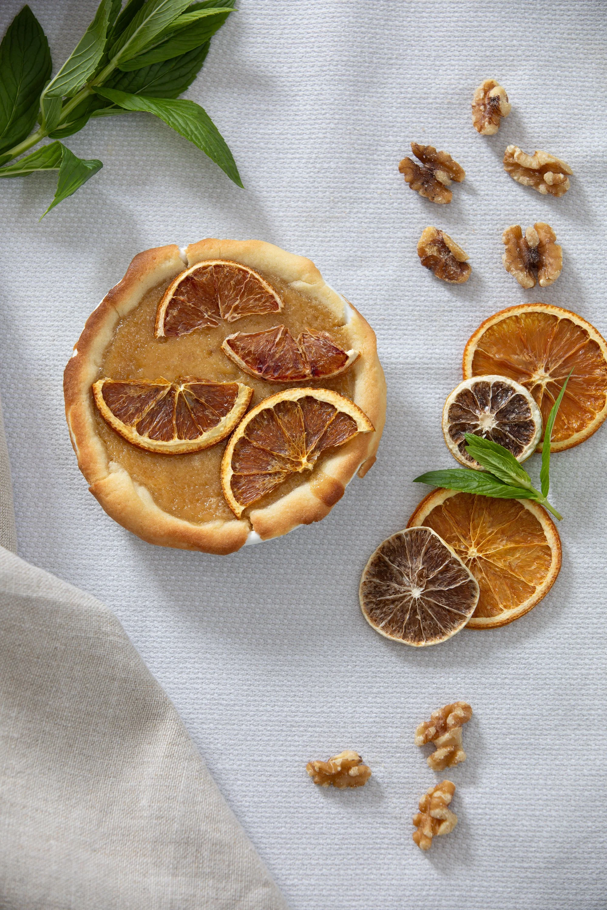 A round lemon tart topped with overlapping dried orange slices, with loose slices of dried orange and some walnuts scattered around on a white textured surface, with sprigs of fresh mint and basil leaves nearby.