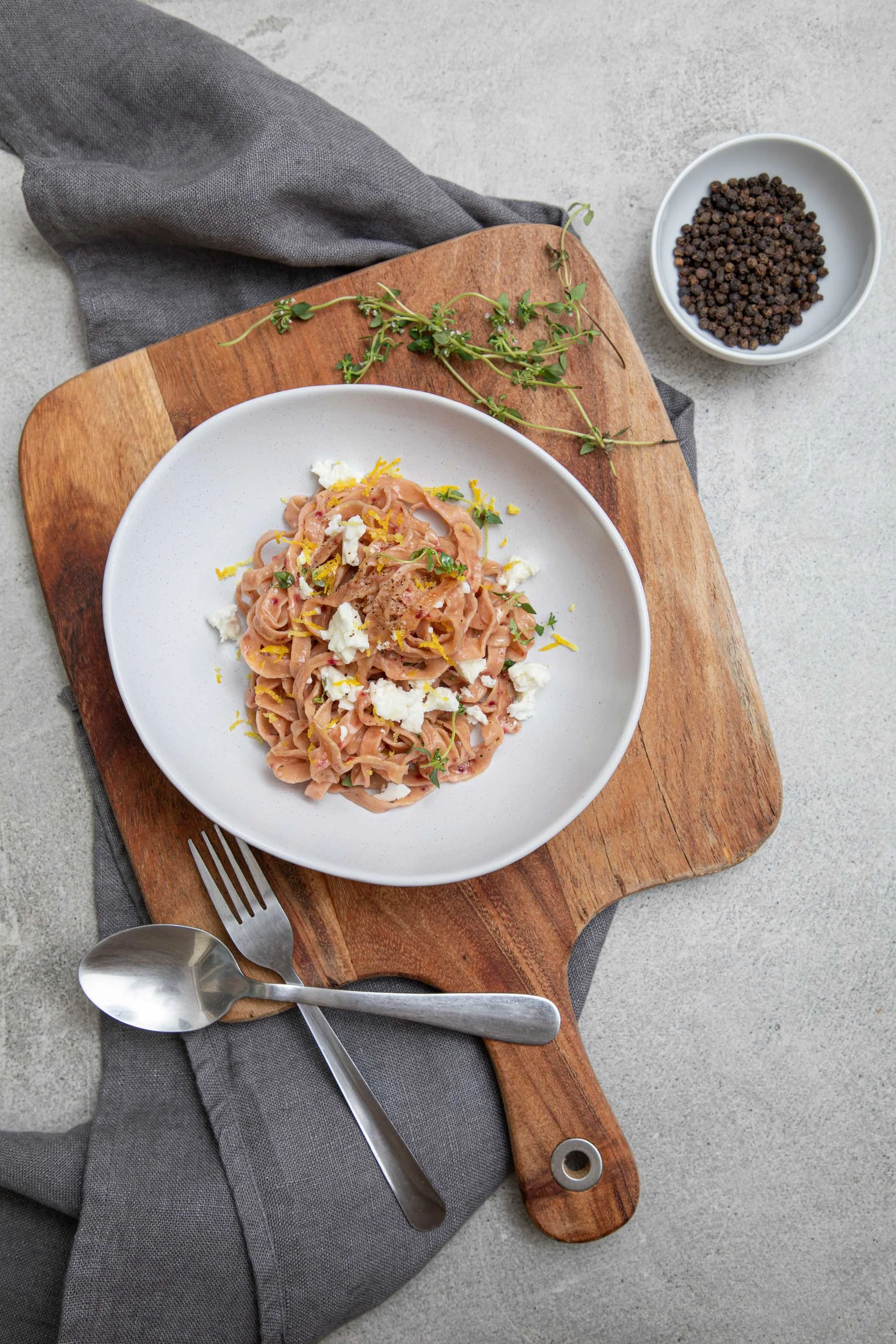 Plate of pasta with herbs, cheese, and lemon zest on a wooden cutting board, with a bowl of peppercorns, a gray napkin, and utensils on a gray surface.