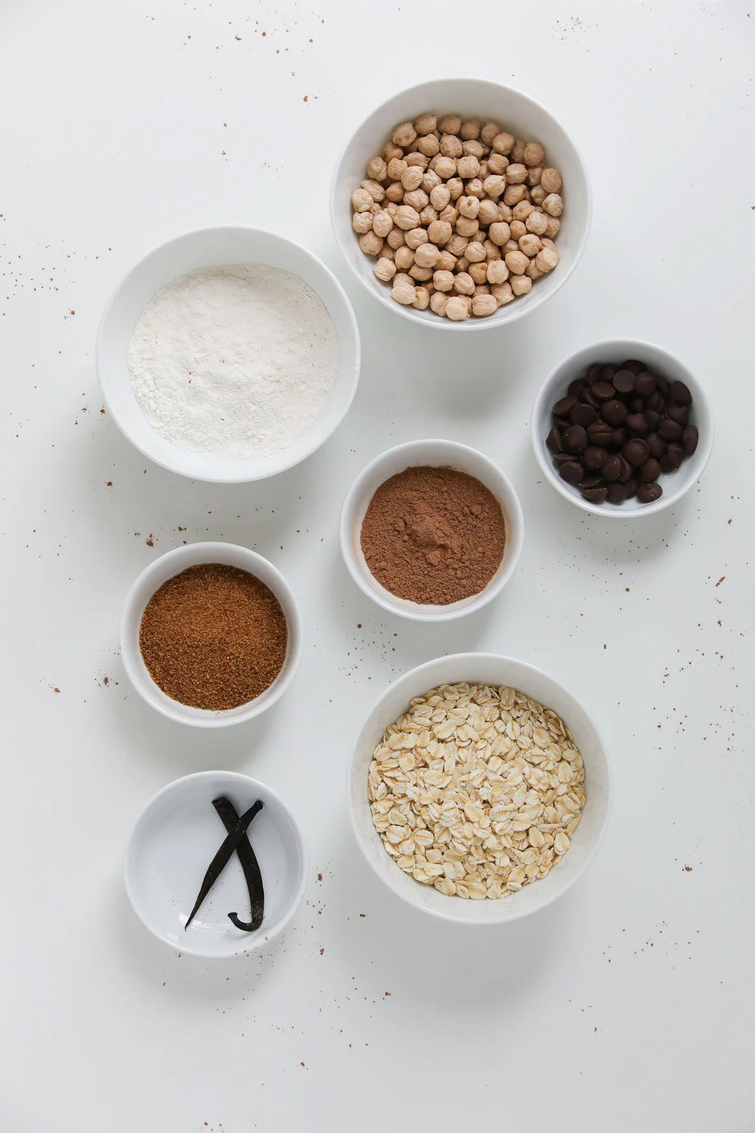Assorted baking ingredients in white bowls on a white surface, including oats, chickpeas, chocolate chips, flour, cocoa powder, spices, and a vanilla bean.