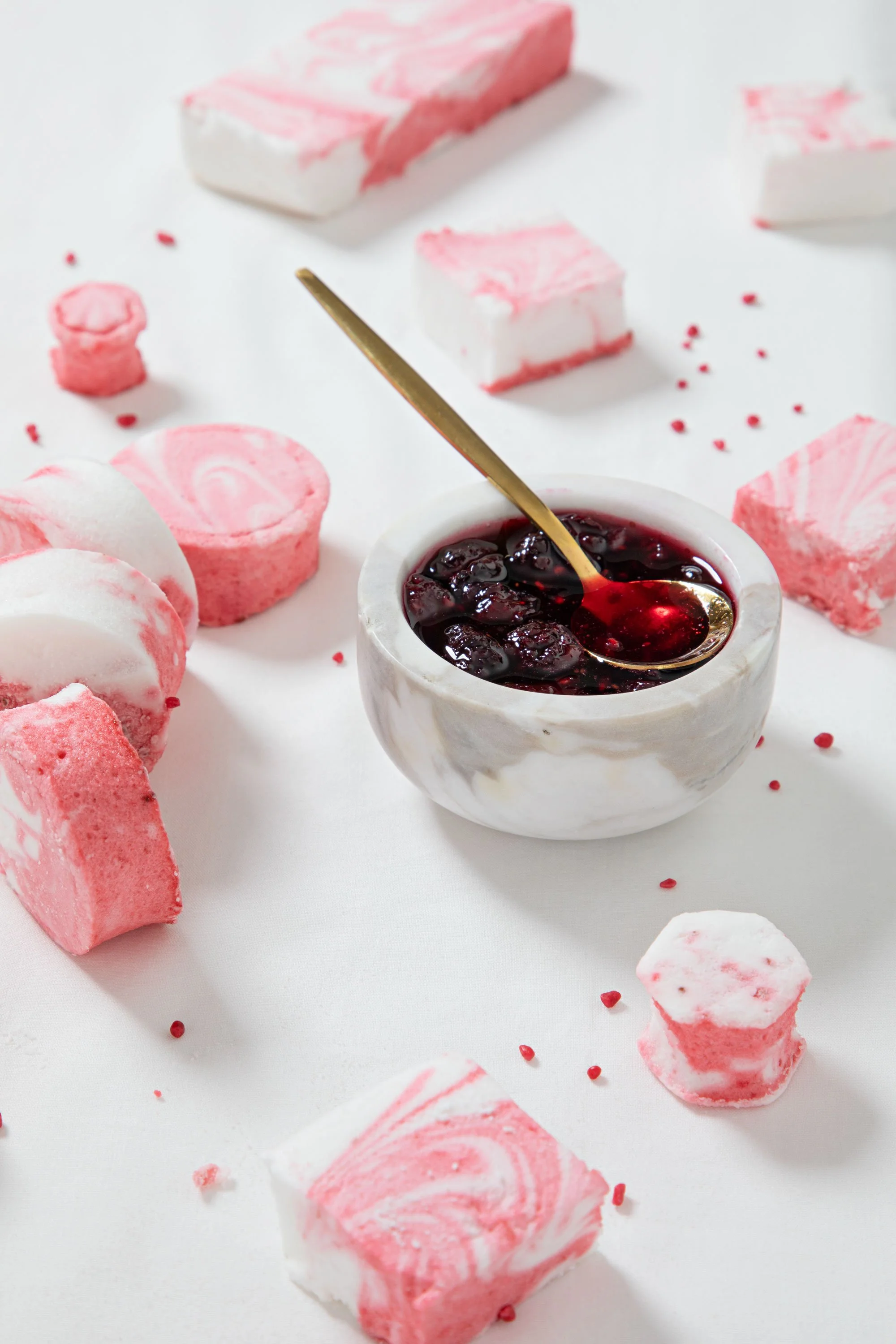 Pink and white marbled soap pieces scattered on a white surface with a small bowl of dark red jam or syrup and a gold spoon.