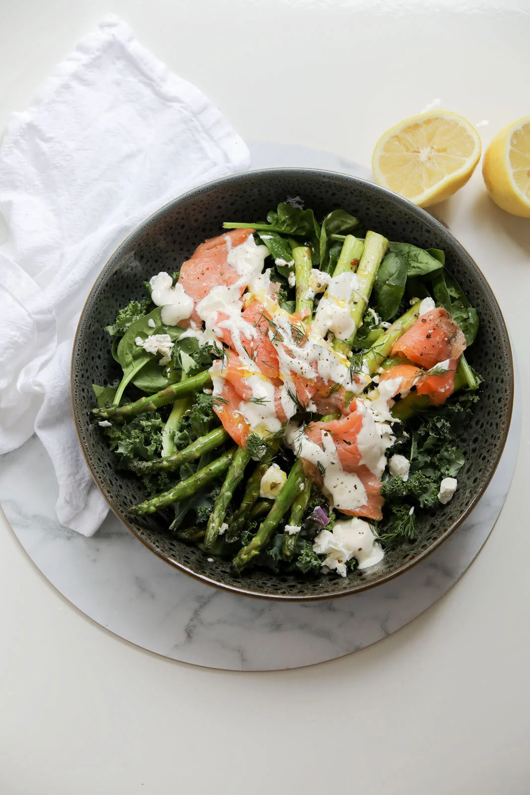 Salad with smoked salmon, goose liver, greens, herbs, and dressing in a dark bowl, with lemon halves and white cloth in the background.