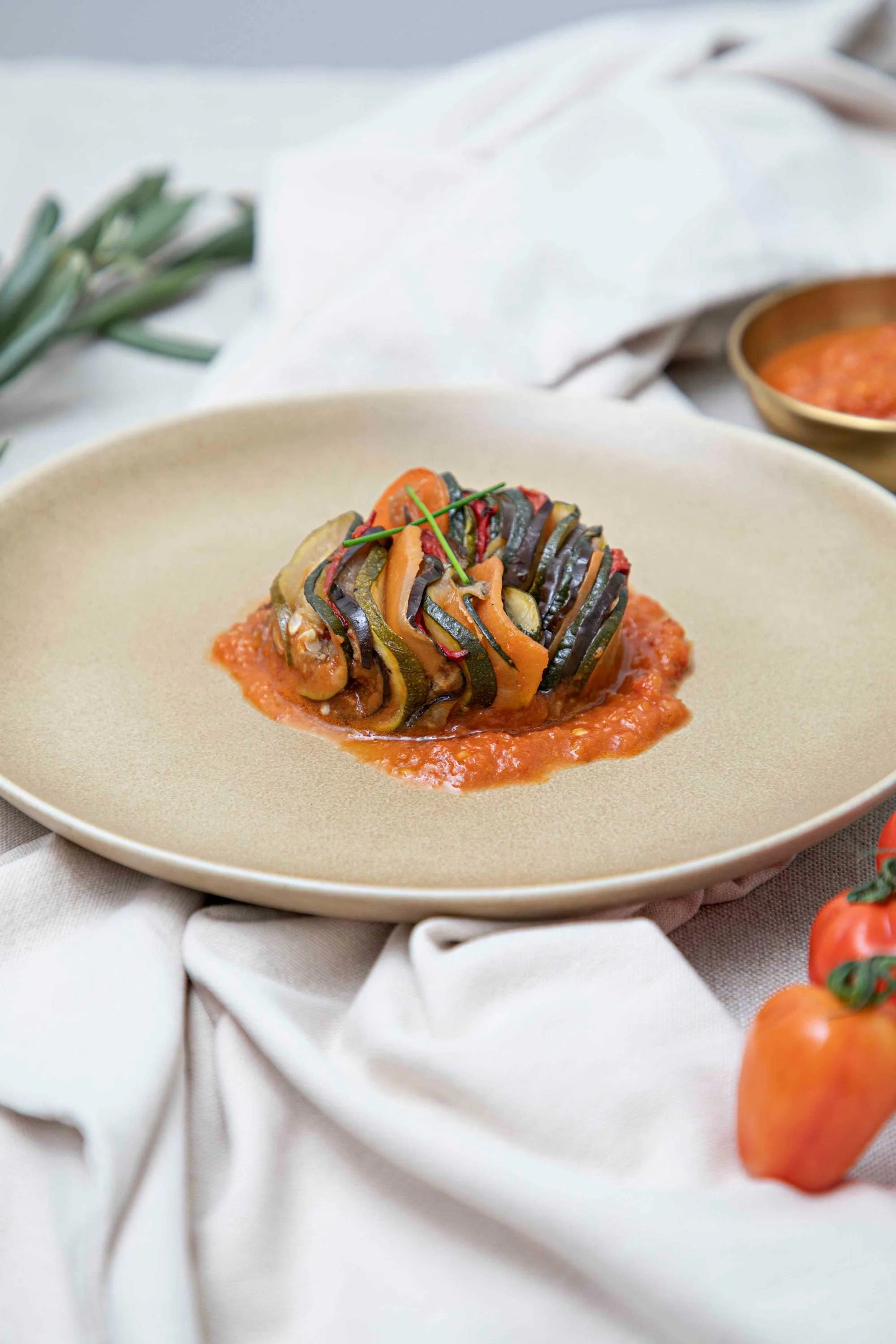 Steamed vegetables, including zucchini and carrots, served on a bed of tomato sauce, garnished with a chive, on a beige plate with a side of tomato sauce in a small bowl, surrounded by a white cloth and fresh produce.