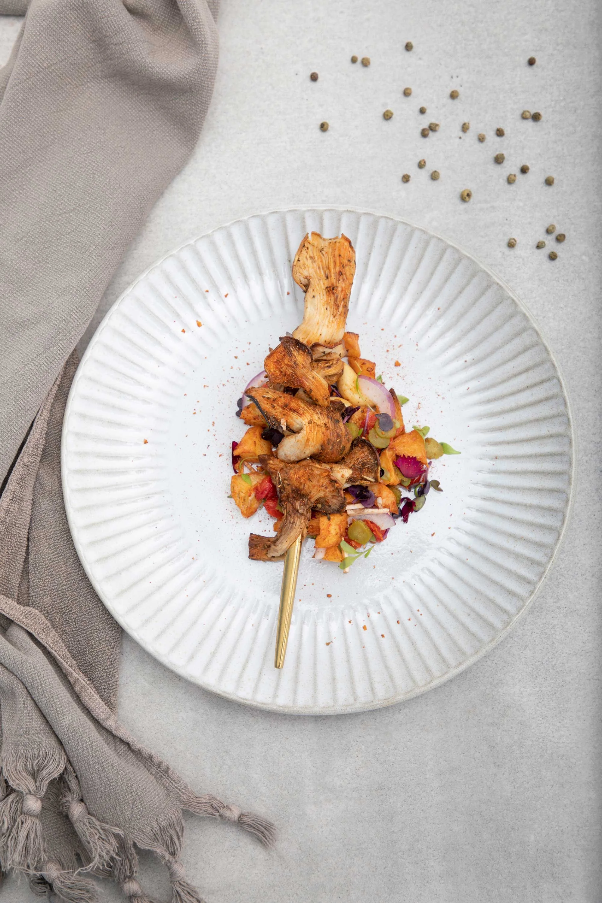 A plated dish with roasted mushrooms and chopped vegetables, garnished with herbs and spices, on a textured white plate, with a beige cloth napkin beside it and scattered peppercorns on the table.
