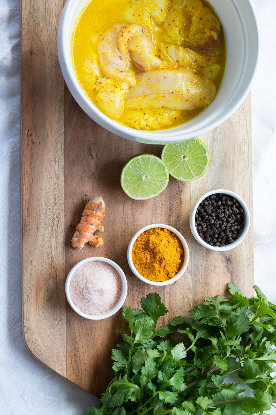 Raw shrimp in a yellow marinade, lime halves, black peppercorns, turmeric powder, pink Himalayan salt, fresh cilantro, ginger root, and black pepper on a wooden cutting board.