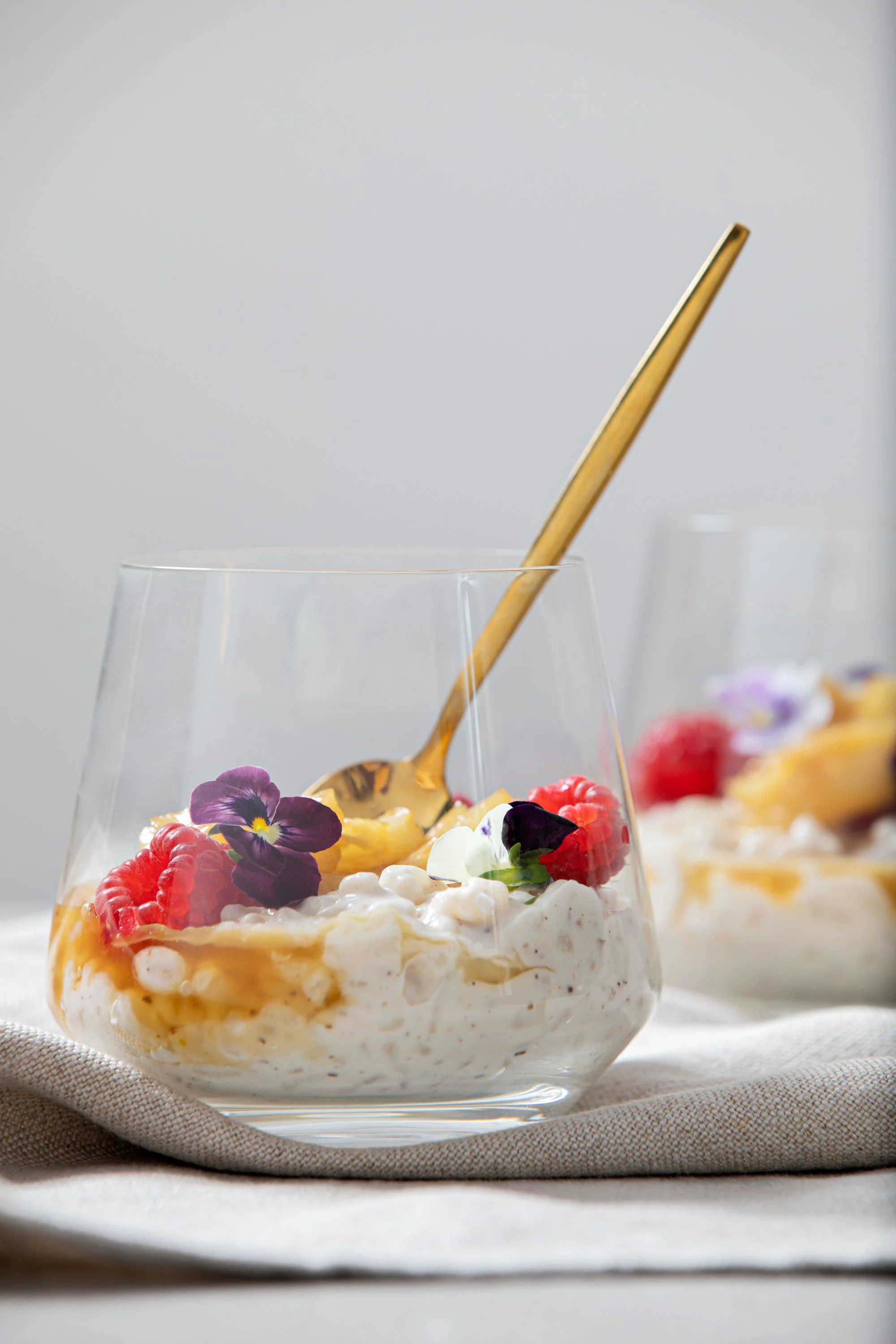 A glass bowl of dessert with whipped cream, raspberries, purple edible flowers, and caramel sauce, with a gold spoon inside.