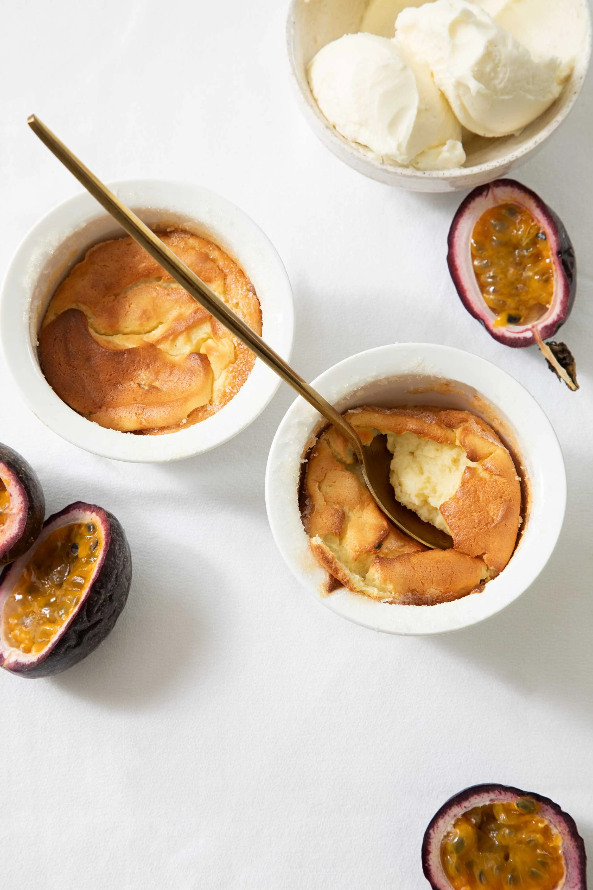 Passion fruits cut in half with bright yellow pulp and black seeds, two small white bowls of vanilla ice cream or frozen yogurt, and two spoons in the bowls on a white surface.