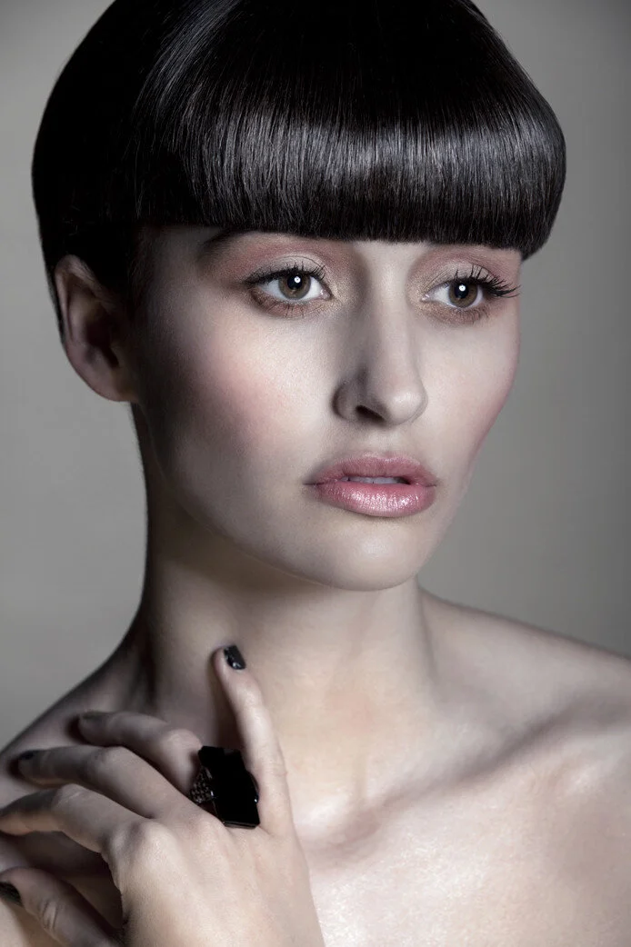 Portrait of a woman with short, dark, straight hair and bangs, wearing makeup, holding a black ring, and looking slightly to the side.