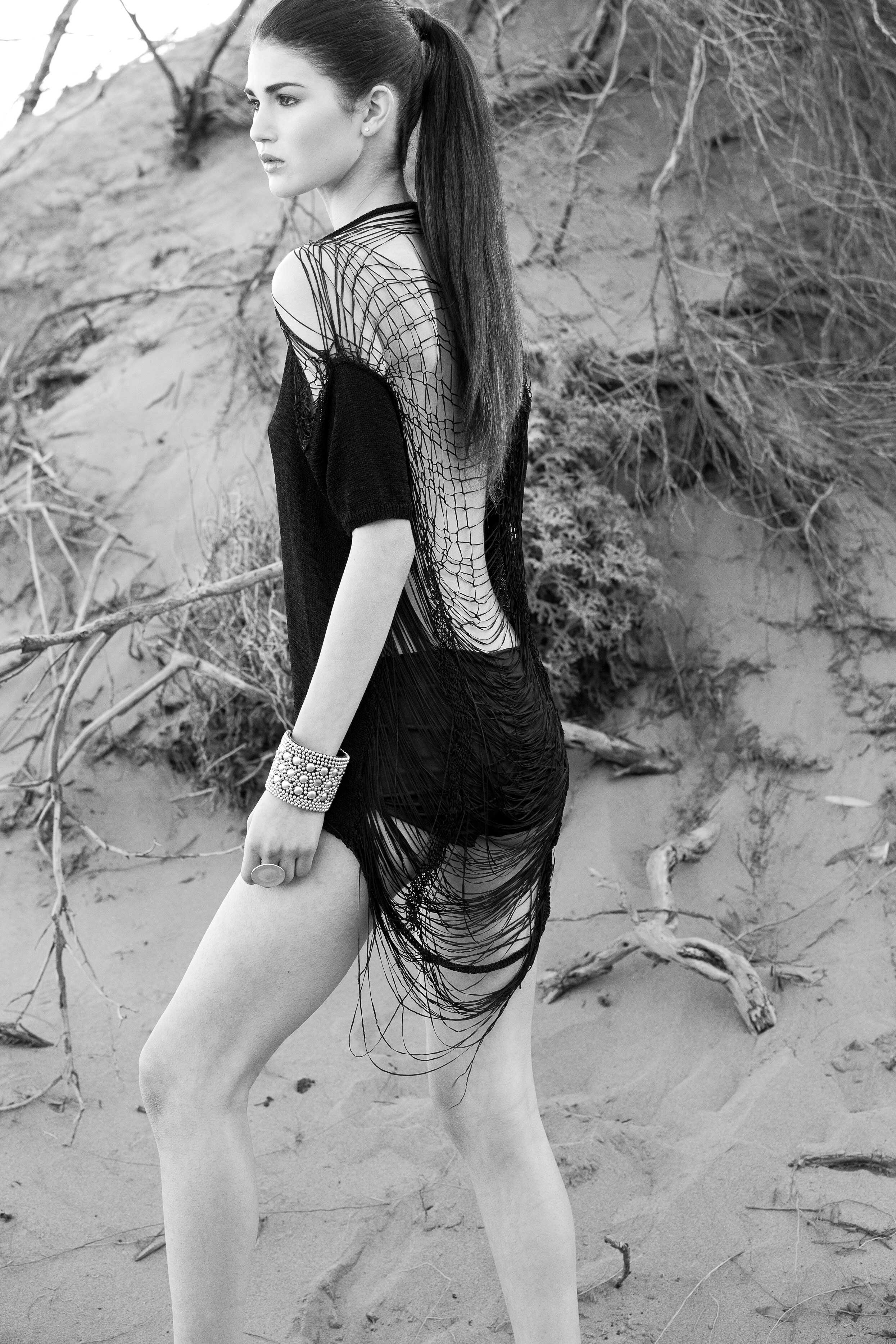 Black and white photo of a woman with long dark hair in a ponytail, standing on a sandy beach with driftwood and dry plants in the background. She is wearing a revealing, see-through, fringed dress and a large bracelet, looking away from the camera.