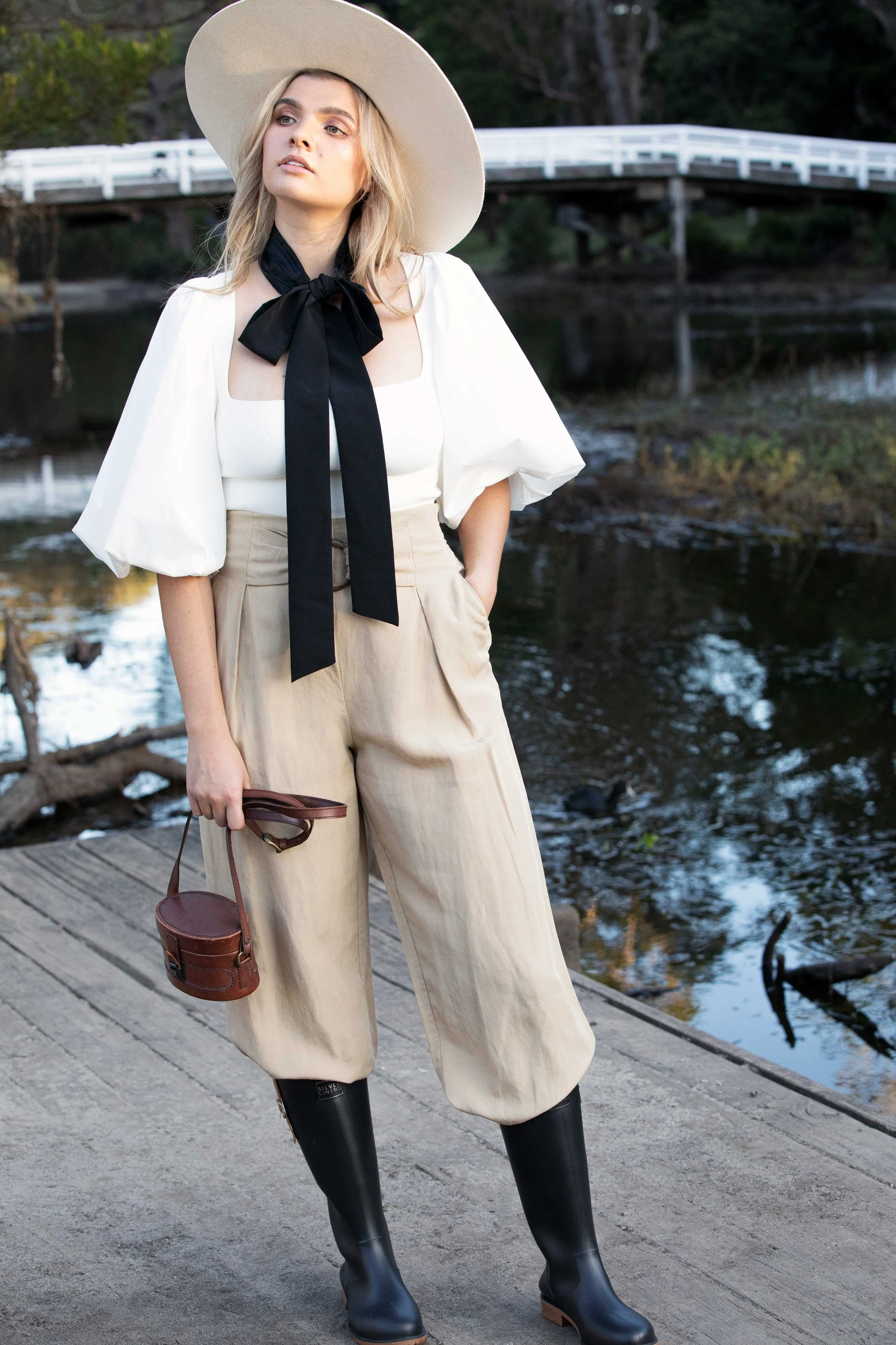A woman standing on a wooden dock near a river, wearing a wide-brimmed hat, white puff-sleeve blouse with black bow tie, beige high-waisted pants, and black rain boots, holding a brown handbag.