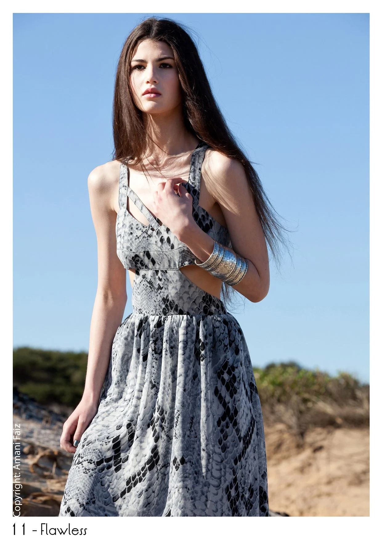 Young woman with long dark hair, wearing a sleeveless snake-print dress, standing outdoors against a clear blue sky with greenery in the background.