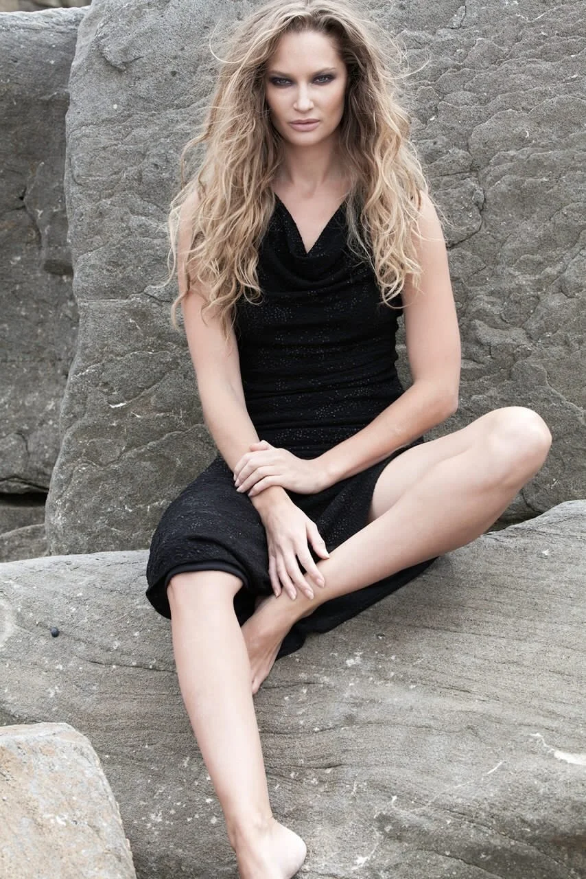 A woman with long, wavy blonde hair sitting barefoot on a large rock in front of a rocky background, wearing a black dress.