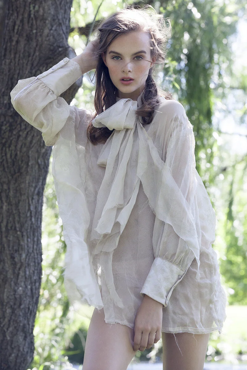 A woman with light skin and long brown hair, wearing a loose, sheer, cream-colored blouse with a large bow at the neckline, posing outdoors near a tree with green foliage in the background.