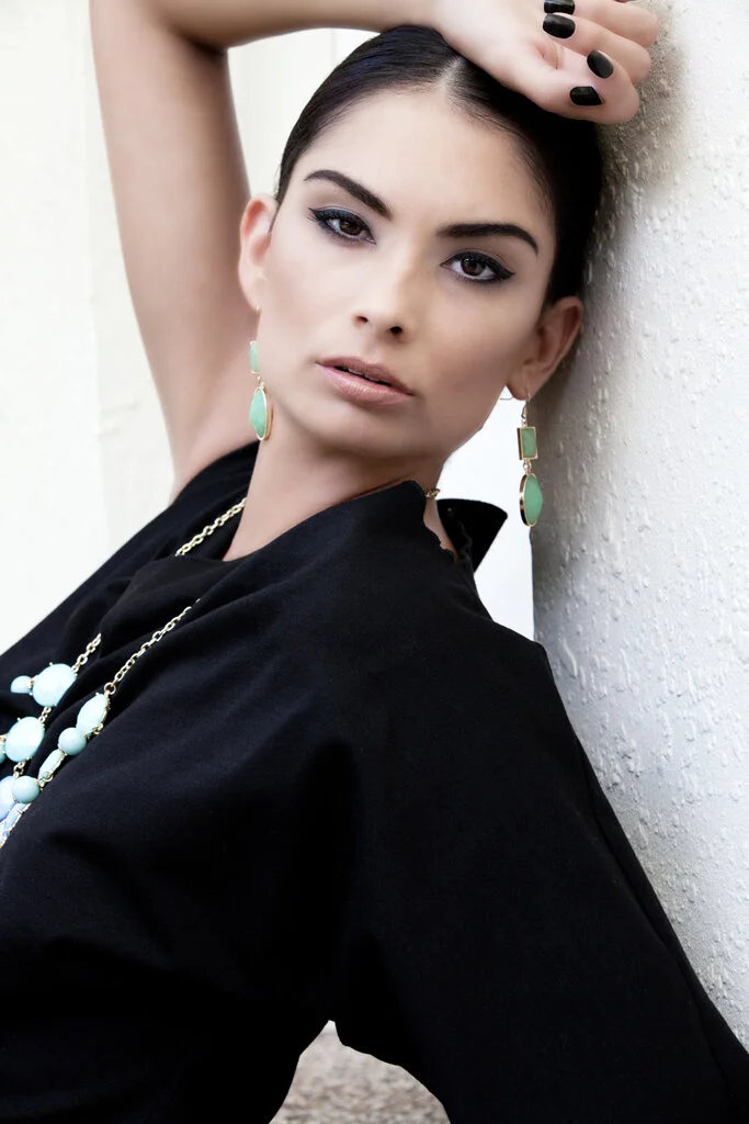 A woman with dark hair and makeup wearing a black top, earrings, and a necklace, leaning against a textured white wall with one arm raised above her head.