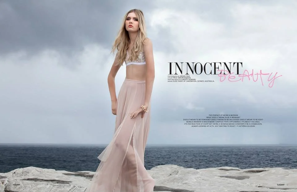 A woman stands on a rocky shoreline with the ocean and cloudy sky in the background, wearing a sheer, flowing peach-colored skirt and a lace crop top.
