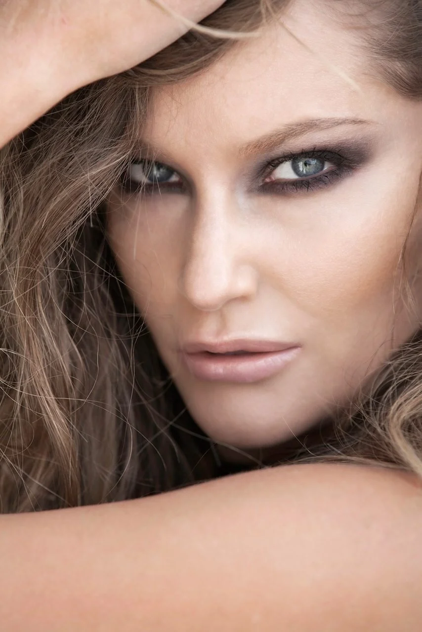 Close-up of a woman with wavy hair, blue eyes, and subtle makeup, looking directly at the camera.