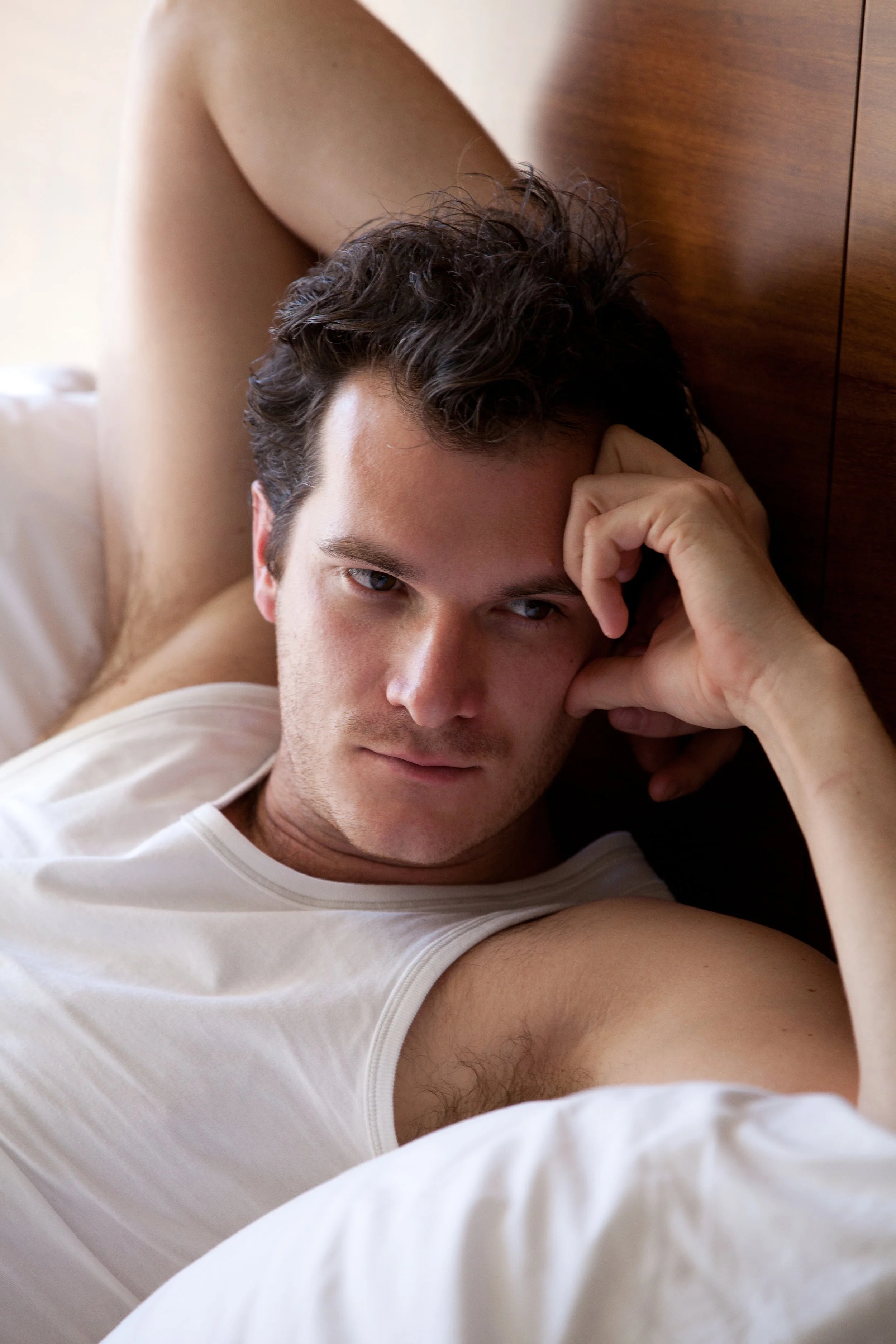 A young man with curly dark hair lying down on a bed with a wooden headboard, wearing a white tank top, gazing into the camera with a relaxed expression, resting his head on his hand.