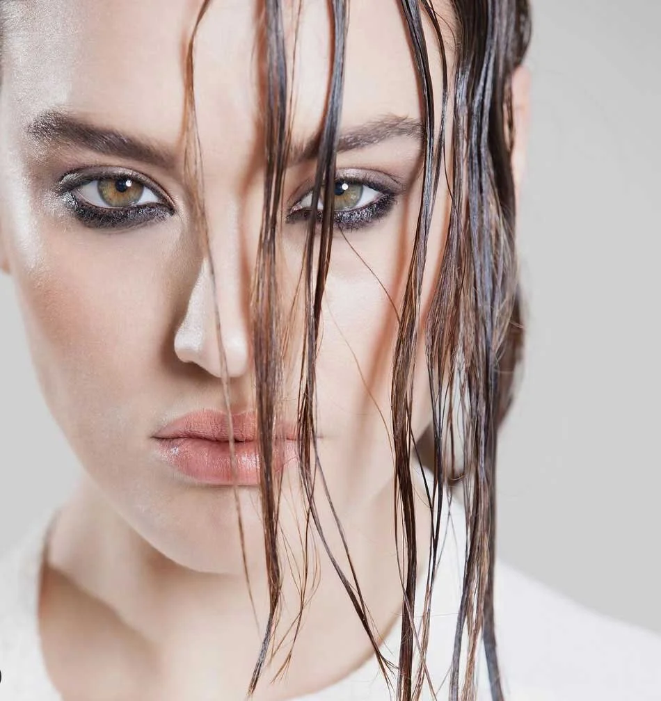 A close-up of a woman with wet hair and makeup, looking directly at the camera.