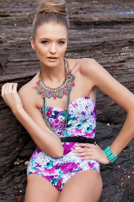 A young woman with blonde hair styled in an elegant updo, wearing a colorful strapless floral swinsuit and multiple layered necklaces. She is sitting outdoors against a rocky background, posing with one hand on her waist and looking at the camera.