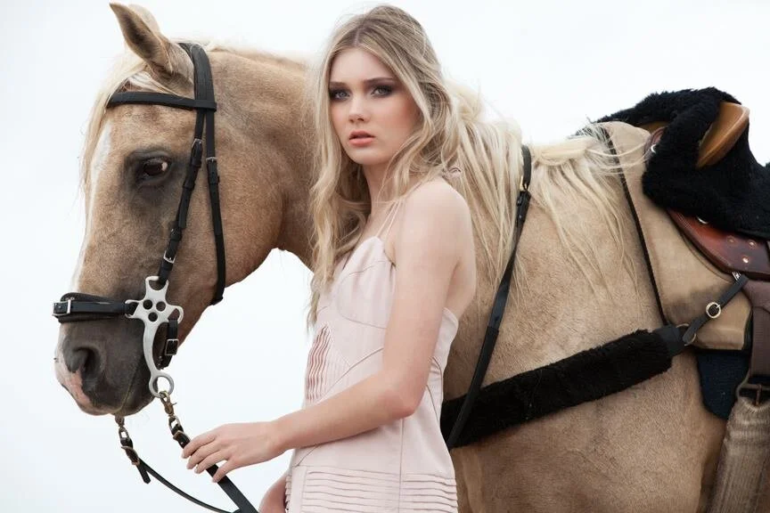 A young woman with long blonde hair standing next to a light-colored horse, holding its reins, with a saddle and riding gear.