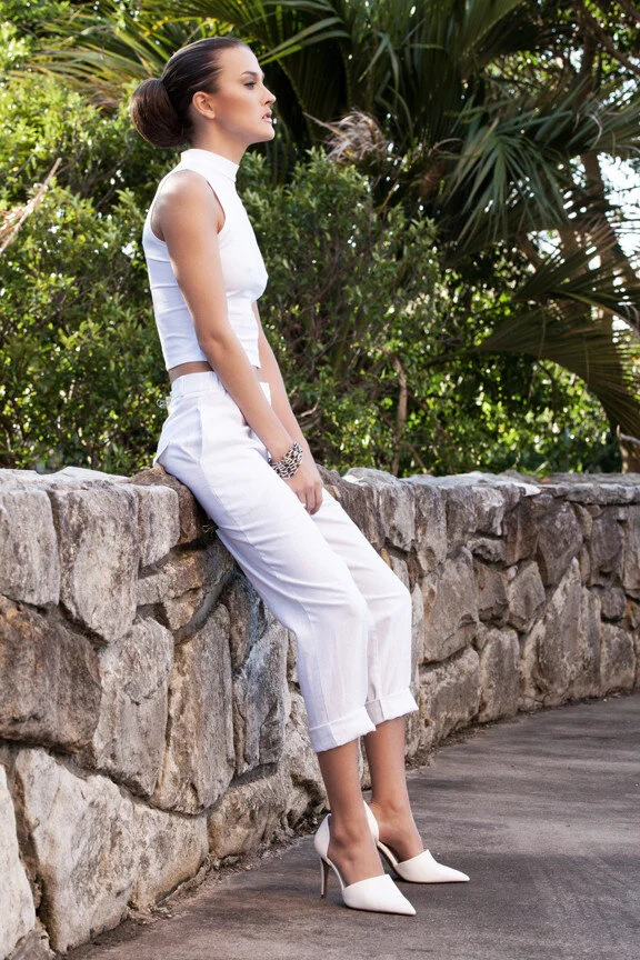 A woman in all white clothing and high heels sitting on a stone ledge outdoors surrounded by lush green plants.