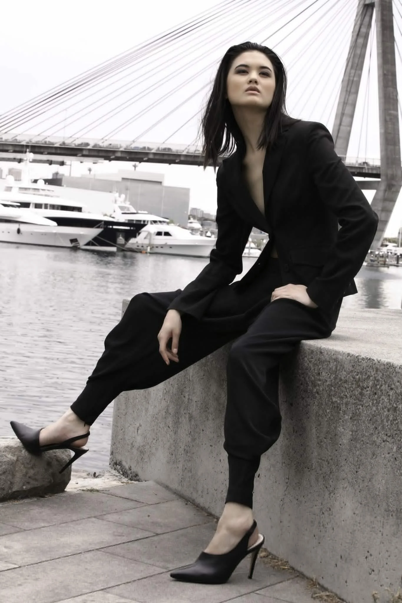 A woman sitting on a concrete ledge near a harbor, wearing a black suit and high heels, with boats and a large bridge in the background.
