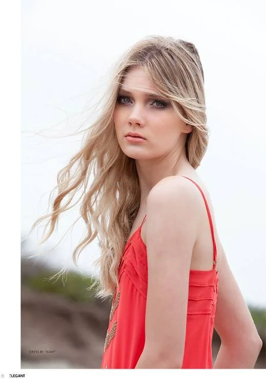 A young woman with long blonde hair and blue eyes wearing a red sleeveless dress on a cloudy outdoor day.