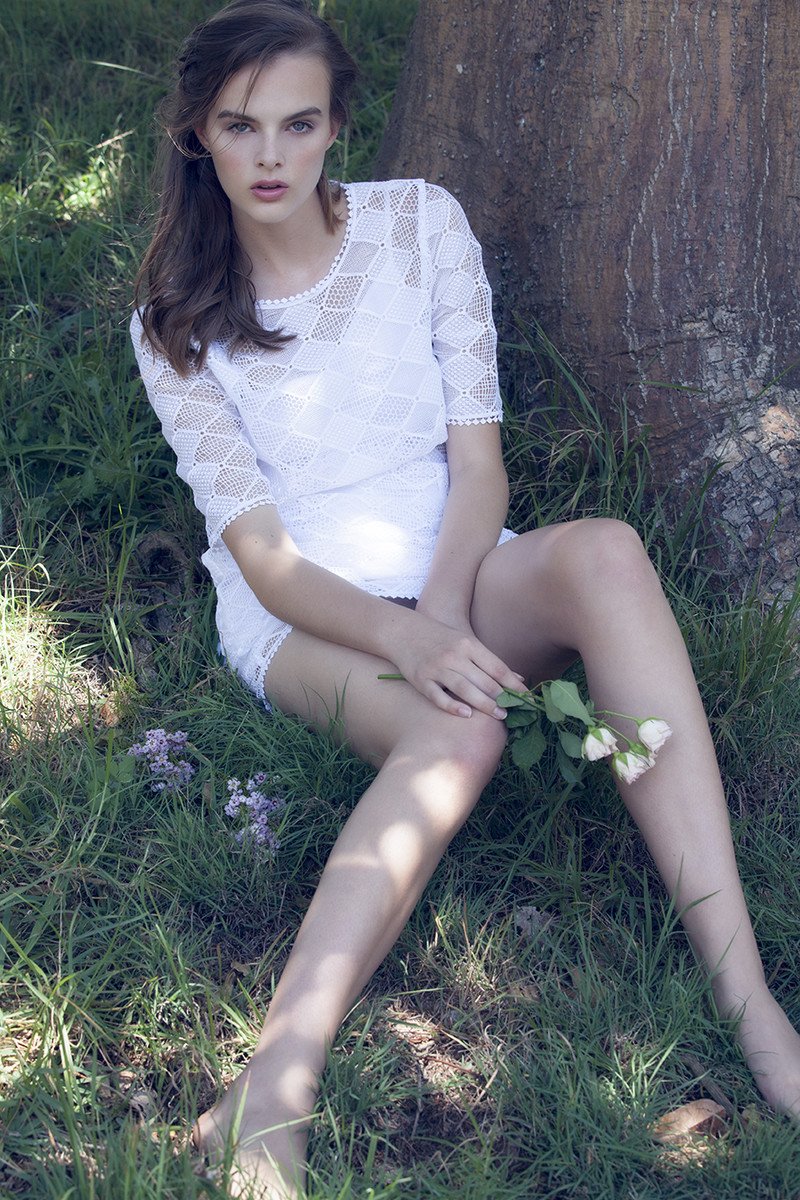 A young woman with long brown hair, wearing a white lace dress, sitting on grass near a tree, holding white roses.