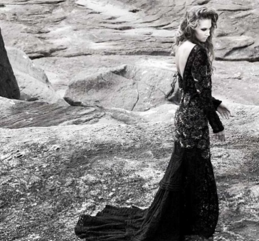A woman with curly hair wearing a long, dark lace dress stands on a rocky terrain with her back partially turned.