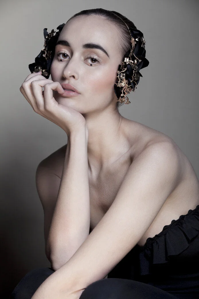 A woman with a decorative headpiece made of black fabric and gold accents, posing with her head resting on her hand, wearing a strapless black off-the-shoulder top, against a neutral background.