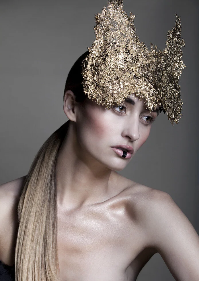 A woman with long blonde hair wears an intricate gold headpiece and makeup with dark eyes and lips.
