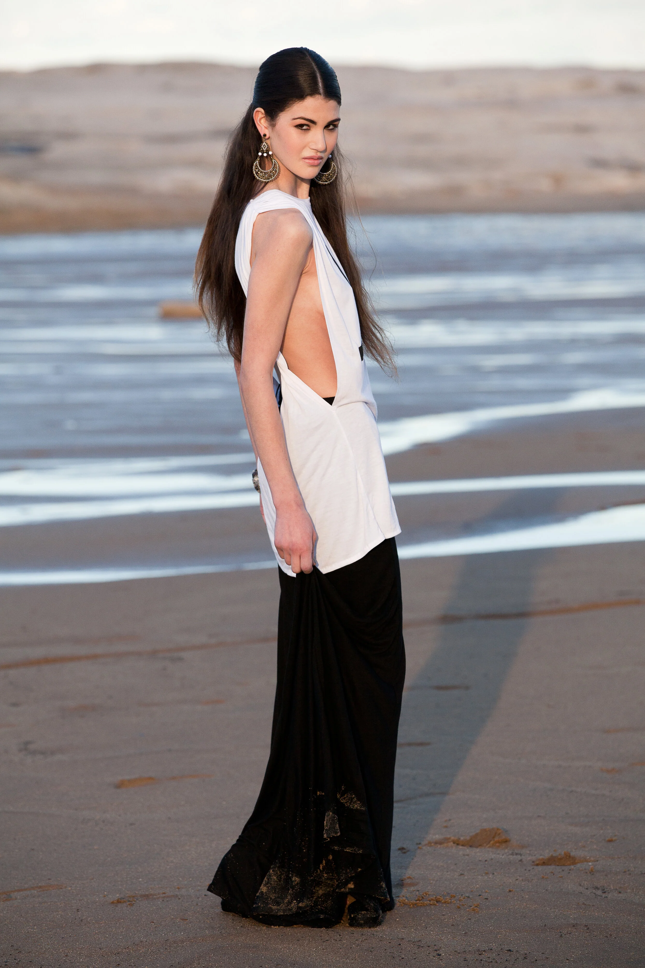 Young woman with long brown hair in a white and black dress standing on a beach near the water with sand and waves in the background.