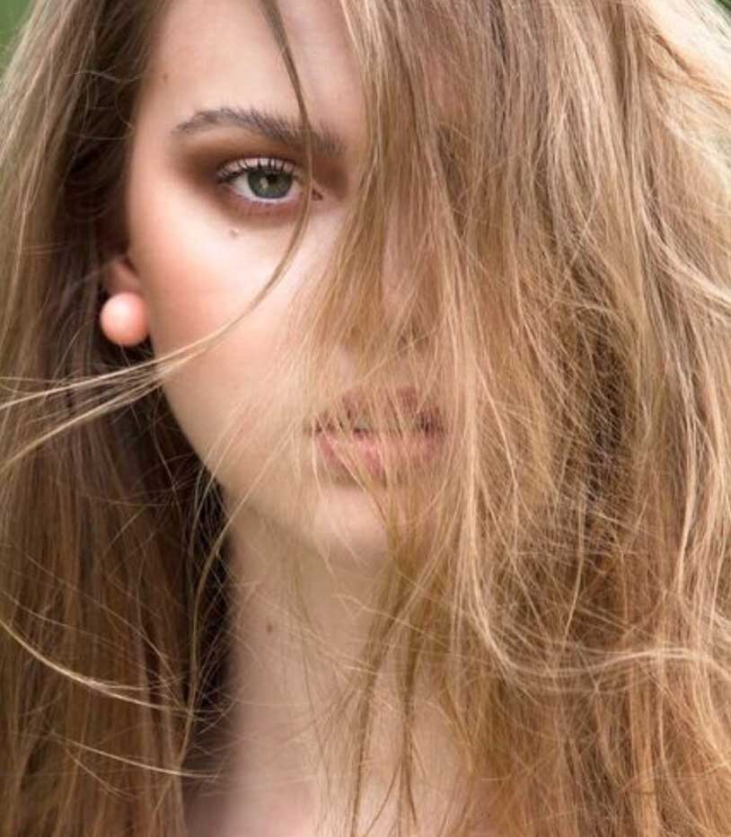 Close-up of a woman's face with tousled, light brown hair partially covering her face, revealing one eye and part of her lips.