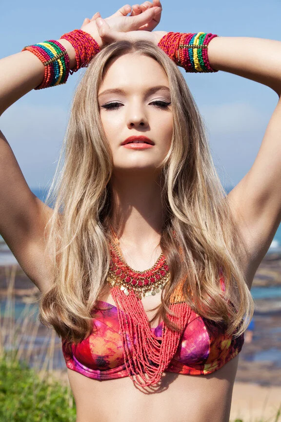 A young woman with long blonde hair is outdoors, wearing colorful beaded bracelets on both wrists, a vibrant patterned top, and layered necklaces. She has her hands raised above her head and is slightly tilting her head with her eyes partially closed