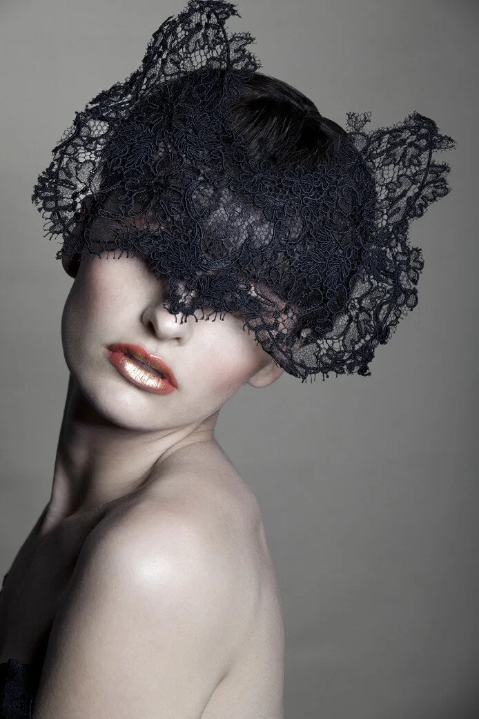 A woman with pale skin wearing a black lace headpiece covering her eyes, lips with metallic gold lipstick, and minimal makeup, against a neutral gray background.