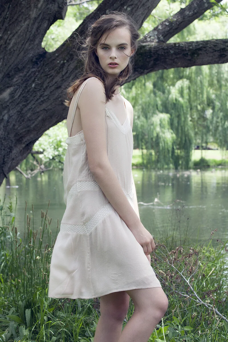 A young woman in a cream-colored dress standing by a tree near a body of water in a lush, green park.
