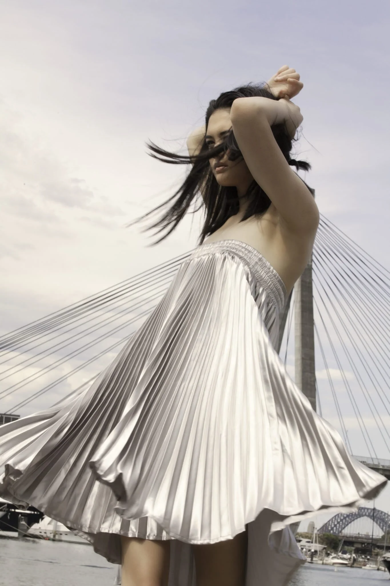 A woman wearing a shiny, pleated, silver dress with a high-low hemline, standing near water with a bridge and cityscape in the background.