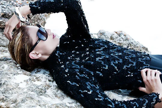 A woman lying on rocks, wearing sunglasses and a black, sequined long-sleeve top.