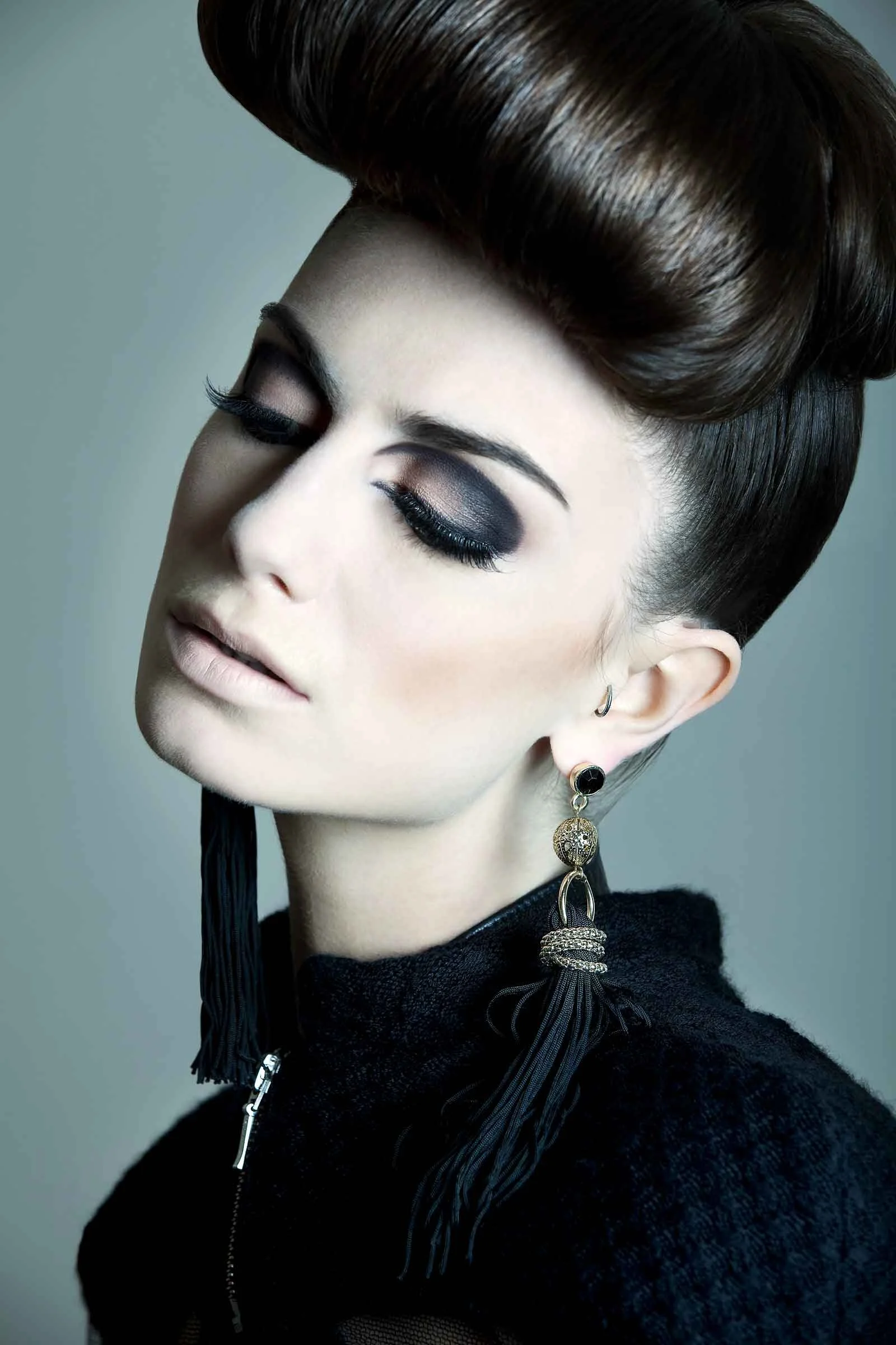 A woman with a vintage hairstyle and makeup, wearing large earrings and a black top with a zipper, posing with her eyes closed against a light background.
