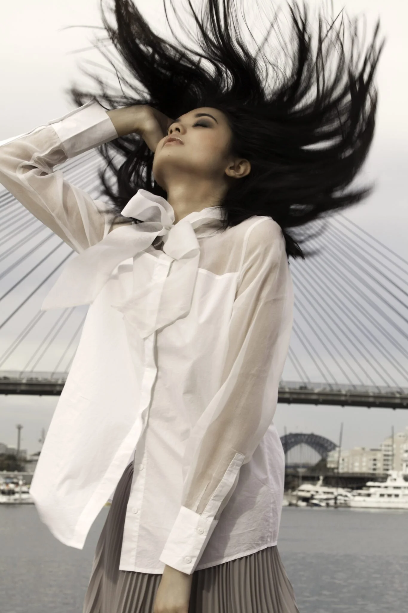 A woman with long black hair, wearing a white blouse and a pleated skirt, stands outdoors with her eyes closed and her hand behind her head. Her hair is flowing upwards in the air, and a bridge and marina with boats are visible in the background.