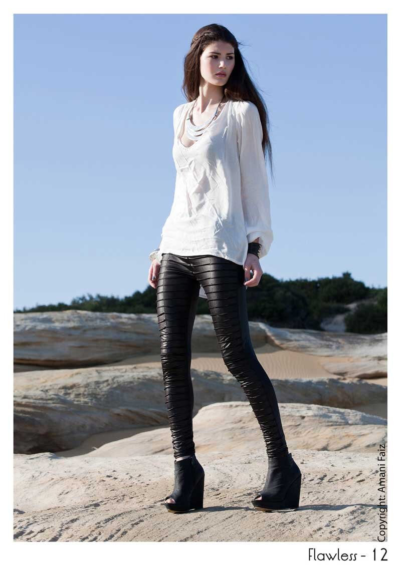 A woman standing outdoors on a sandy surface with rocks and sparse vegetation, wearing a white blouse, black leather pants, and black open-toe high-heeled boots, with a blue sky background.