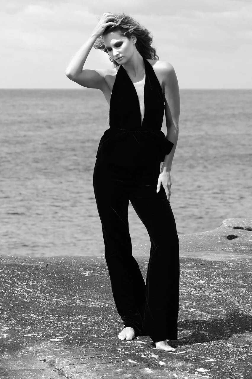 Black and white photo of a woman in a sleeveless, deep V-neck outfit standing barefoot on a rocky shoreline by the ocean, looking down with one hand on her head.