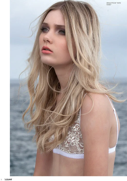 A young woman with long, wavy blonde hair and fair skin standing outdoors near a body of water, looking off into the distance with a thoughtful expression.