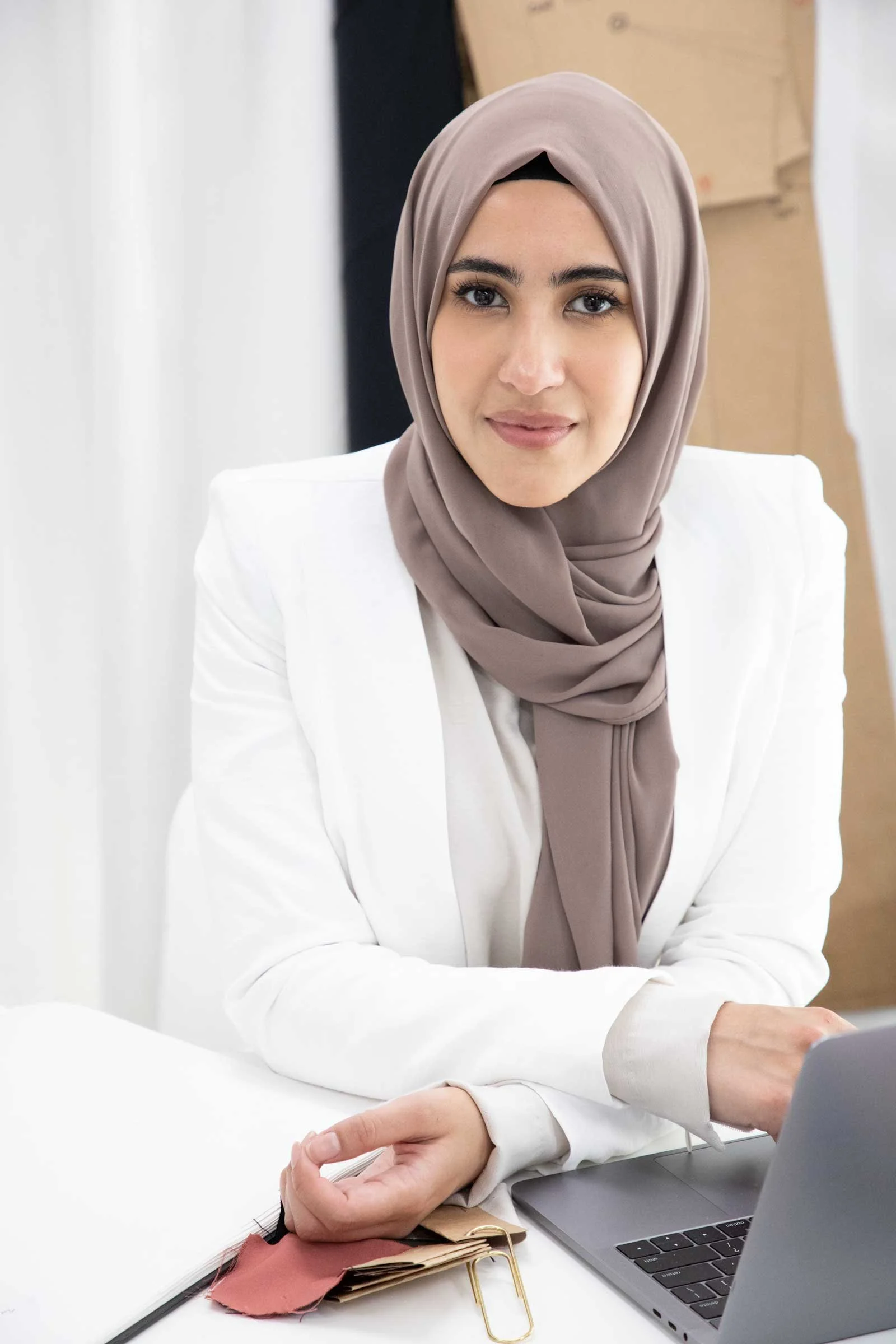 A woman wearing a beige hijab and white blazer sitting at a desk with a laptop, color swatches, and fabric samples.