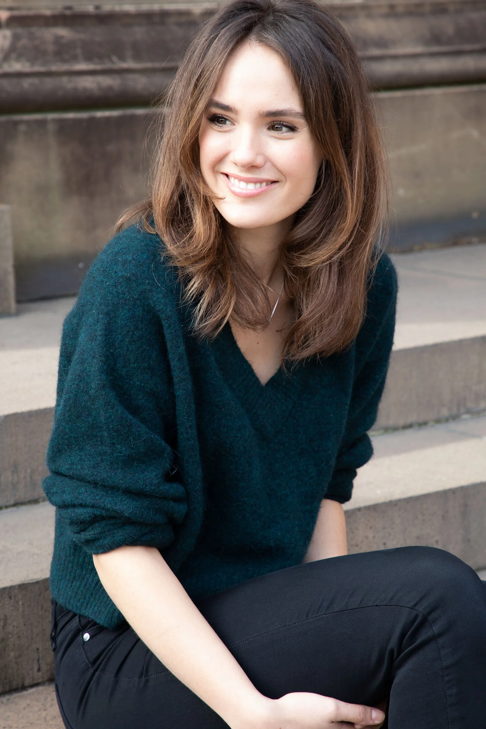 A young woman with long brown hair and a bright smile sitting on outdoor steps, wearing a dark teal sweater and black pants.