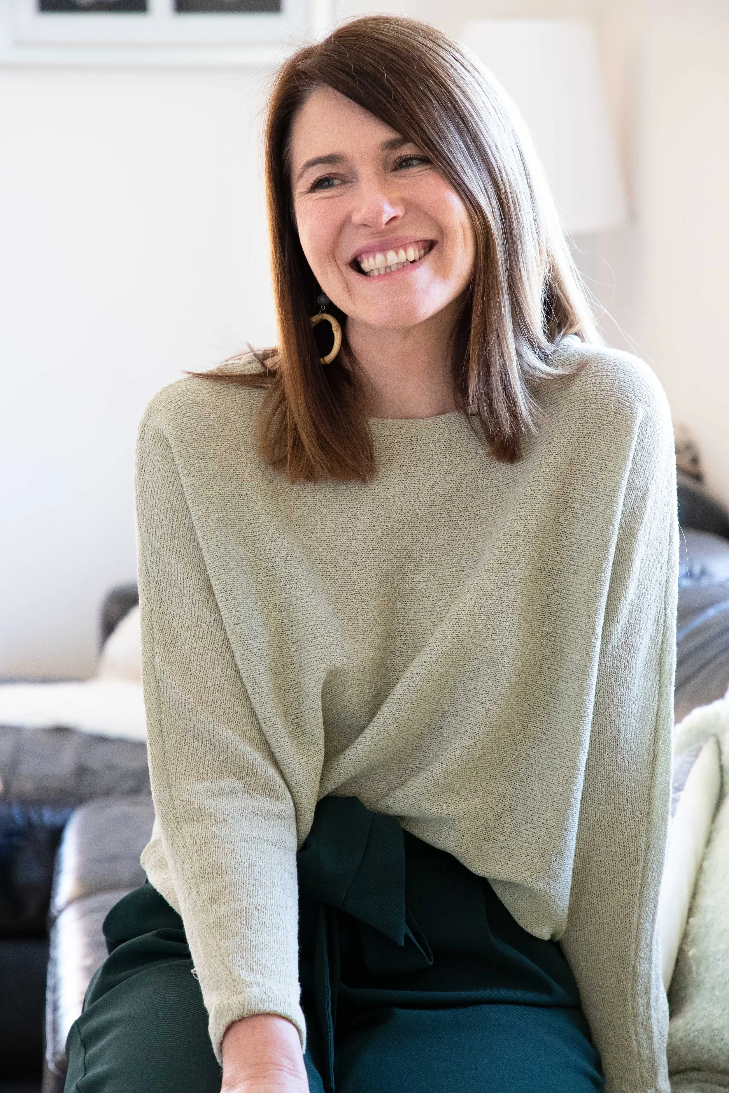 A woman with shoulder-length brown hair, smiling and wearing a beige sweater with large earrings, sitting on a couch in a bright room.