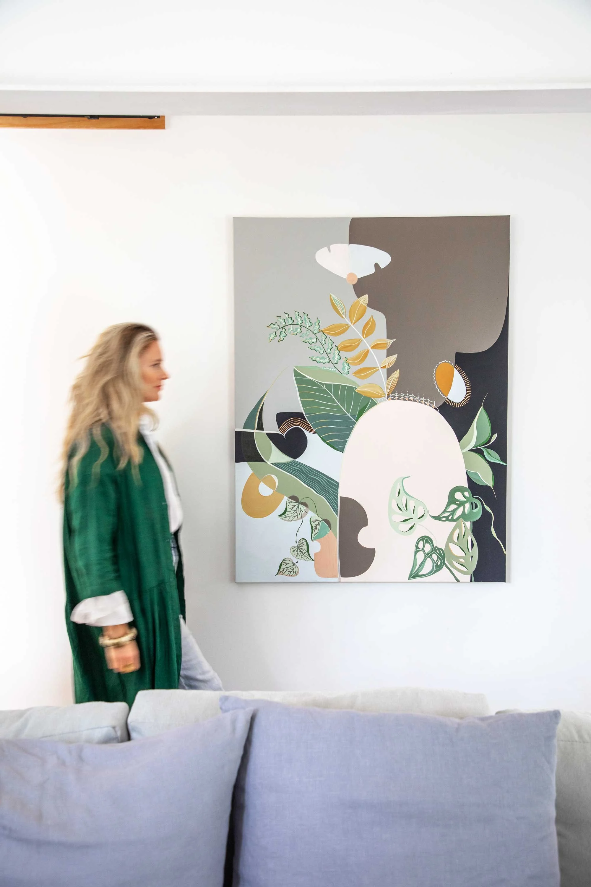 A woman with long blonde hair wearing a green jacket and white pants walking past a large abstract painting of a face made of various plant and leaf patterns in a white-walled gallery.